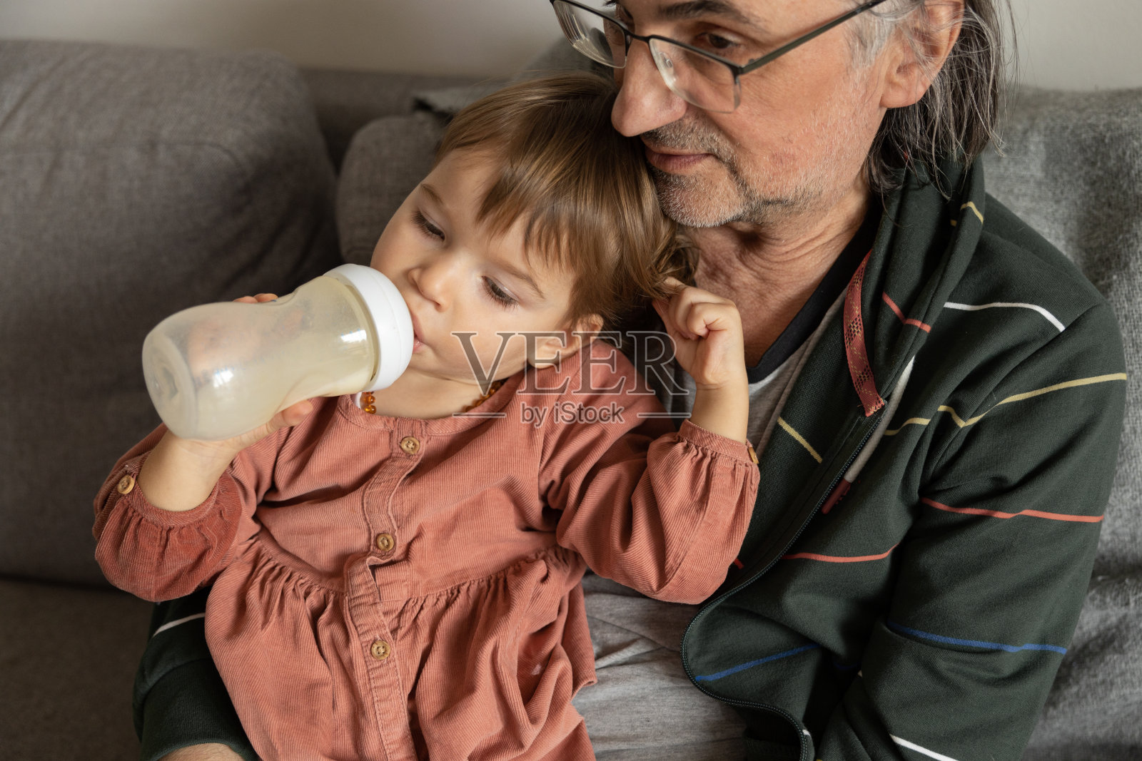 一个小女孩坐在家里的沙发上，坐在爷爷旁边，喝着瓶子里的牛奶。照片摄影图片