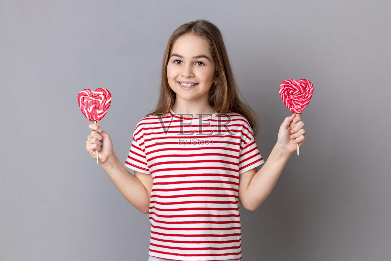 小女孩可口的心形棒棒糖，表情俏皮，微笑着看着镜头。照片摄影图片
