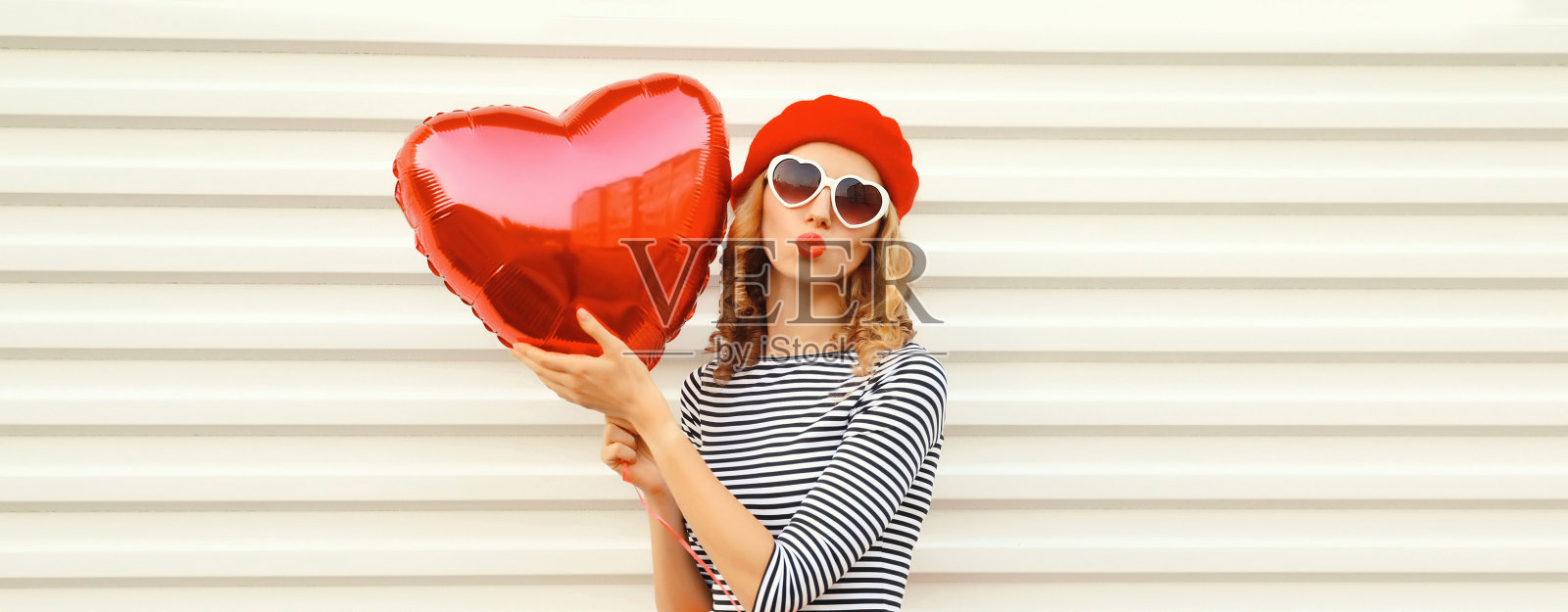 快乐的年轻女子的肖像与红色心形气球吹她的嘴唇送甜蜜的飞吻戴贝雷帽在白色背景照片摄影图片