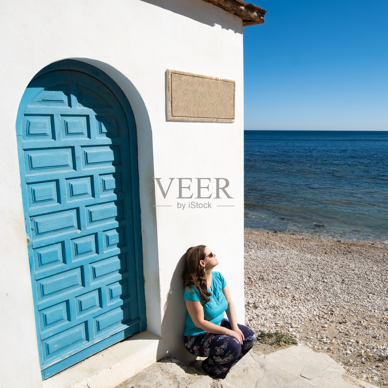 在一个阳光明媚、温暖的冬日早晨，一个金发的中年白人妇女弯着腰靠在一堵有蓝色门的白墙上，在海边晒日光浴。广场组成。Portitxol湾，爪哇。照片摄影图片