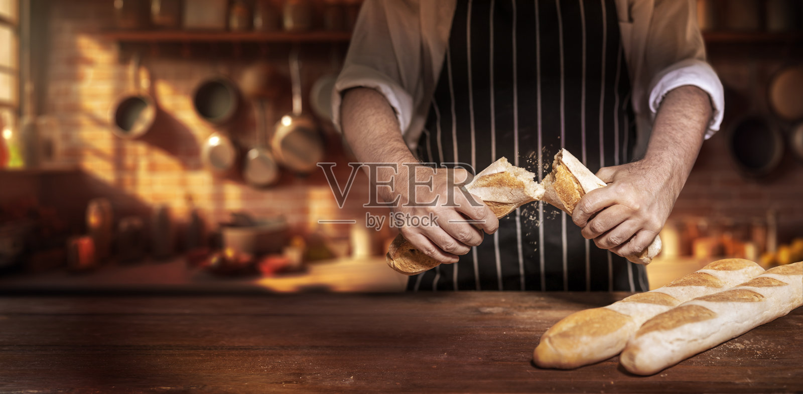 厨师在老式厨房的木桌上把长棍面包掰成两半照片摄影图片