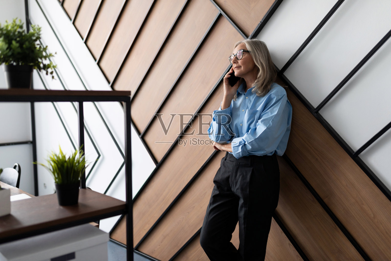 50岁的成功商业女性高管在办公室靠在墙上讲手机。专业女会计师做会计财务发票的概念照片摄影图片