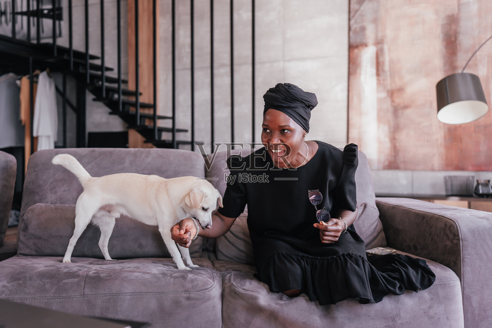 年轻的成年非洲妇女在黑色头巾和裙子玩狗坐在家里的沙发上笑着玩得很开心。白色小狗咬伤了主人的手在酒店房间与非洲裔美国女孩玩耍。调皮的宠物。照片摄影图片