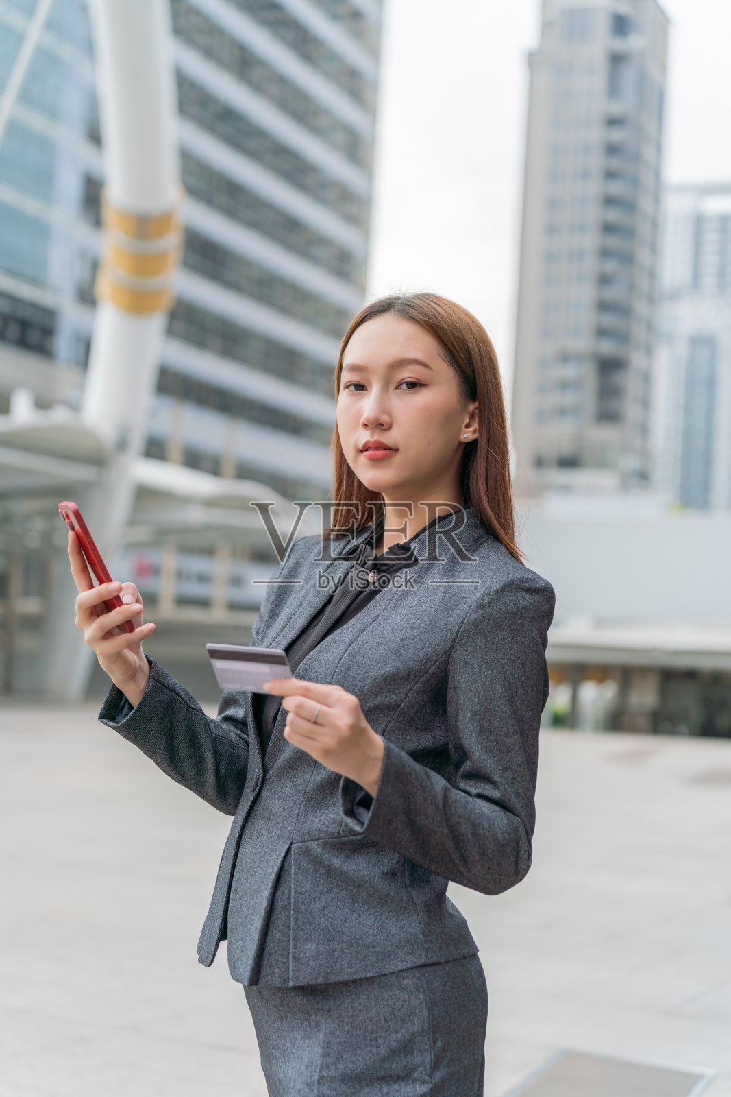 一名年轻的亚洲女商人穿着灰色的办公套装，在商业区用她的智能手机使用信用卡进行在线交易，她微笑着自信照片摄影图片