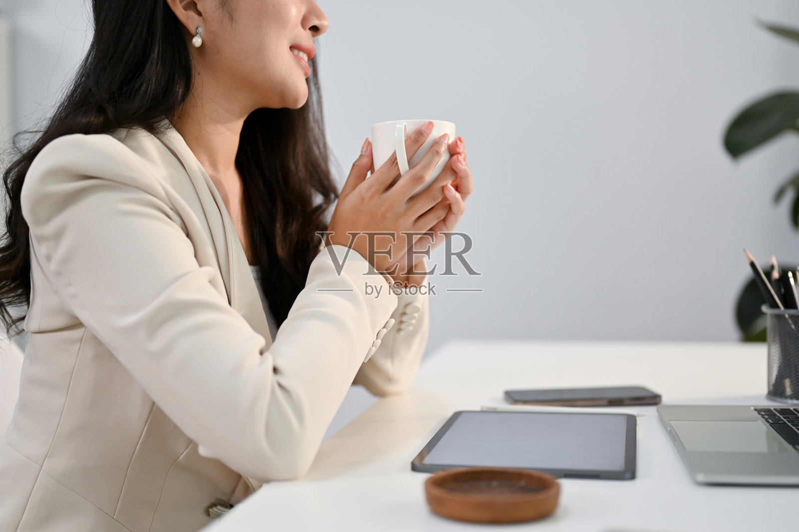 一个放松的亚洲女商人坐在办公桌前拿着咖啡杯的剪裁图片。照片摄影图片