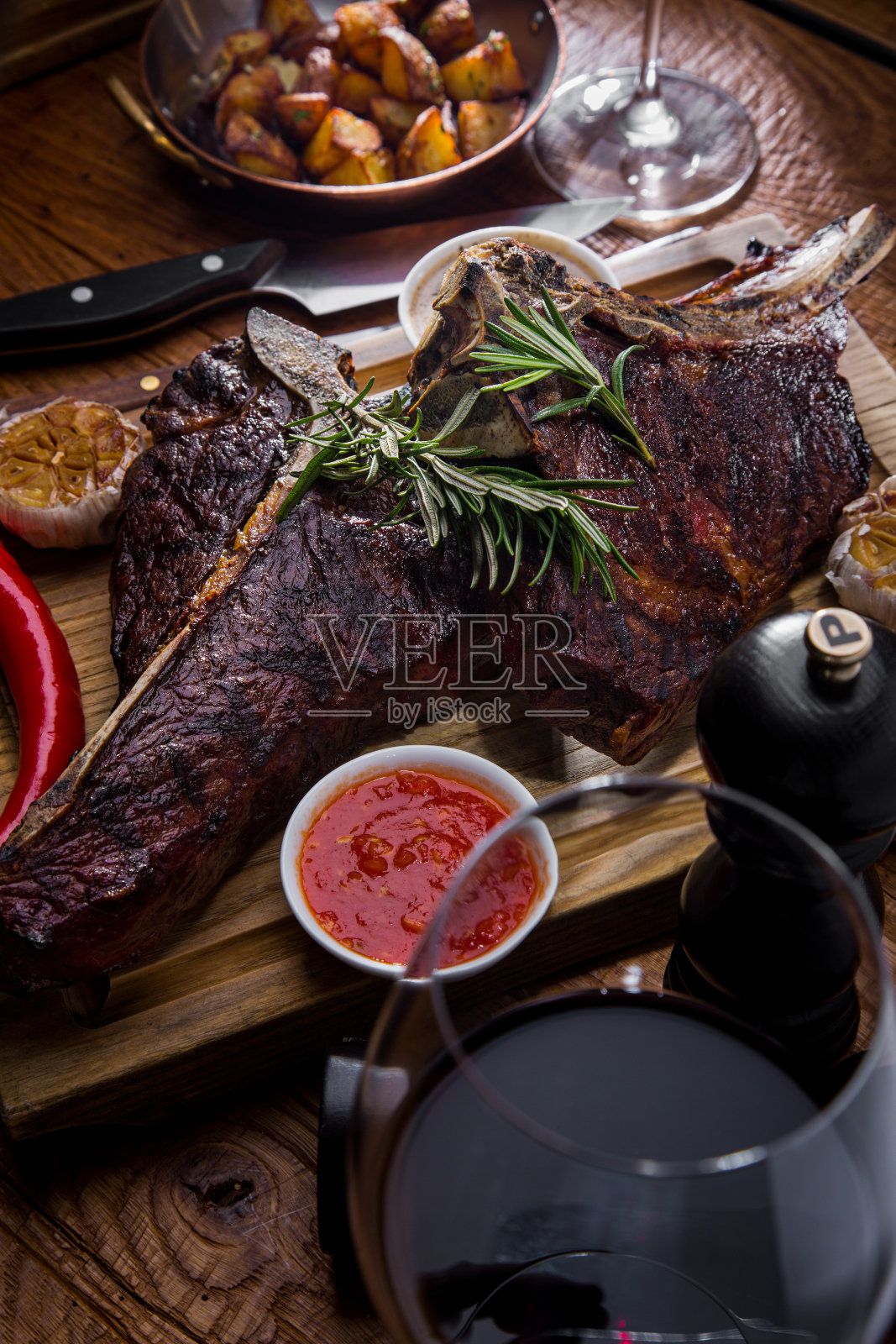 用香料、大蒜、胡椒粉、迷迭香、红白酱汁烤牛排，用木板煎土豆，配餐具，红酒酒杯，胡椒粉瓶和盐瓶。垂直方向照片摄影图片