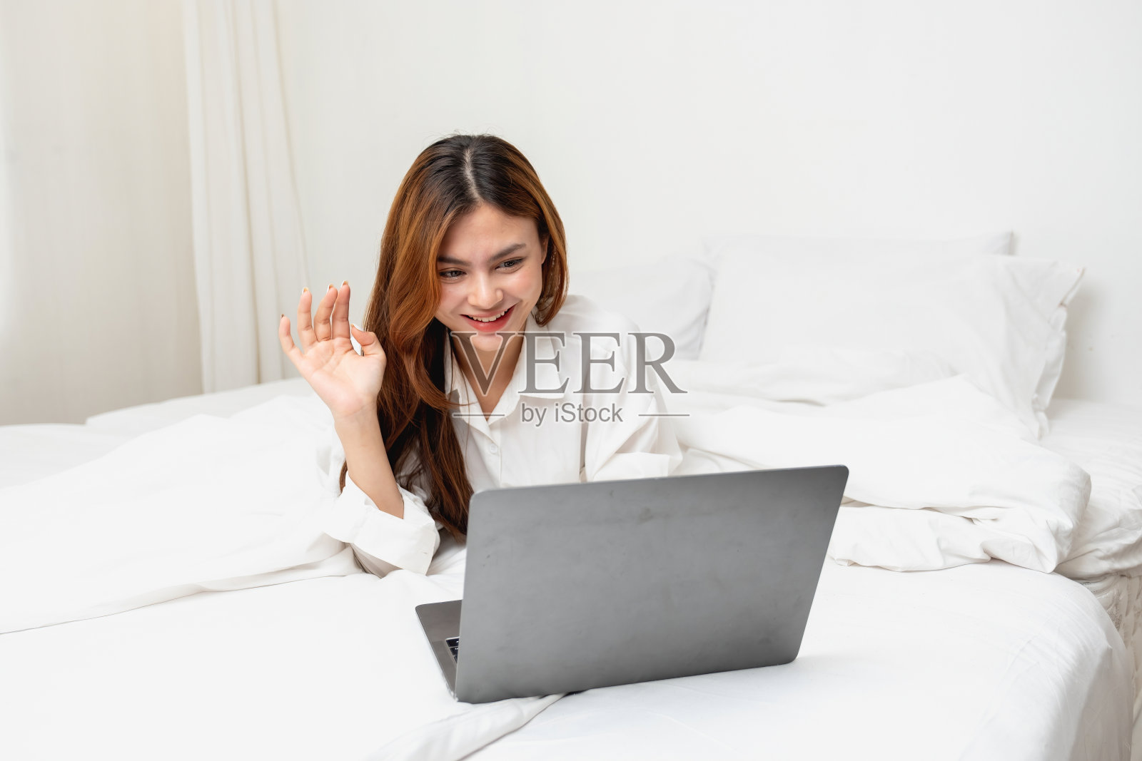 穿着白色睡衣的女人在周末早上醒来，休息放松，玩笔记本电脑手机，在白色卧室的玻璃里吃面包和喝茶。早上度假的概念。照片摄影图片