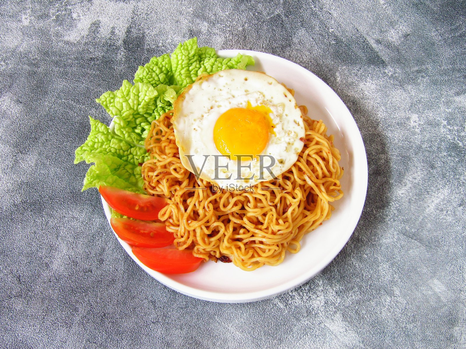 Mie Sedap Goreng，是一种印尼即时炒面，除了Indomie Goreng外，它也很受欢迎。盛在白色盘子里，鸡蛋、西红柿和白菜朝上。在深色背景上。照片摄影图片