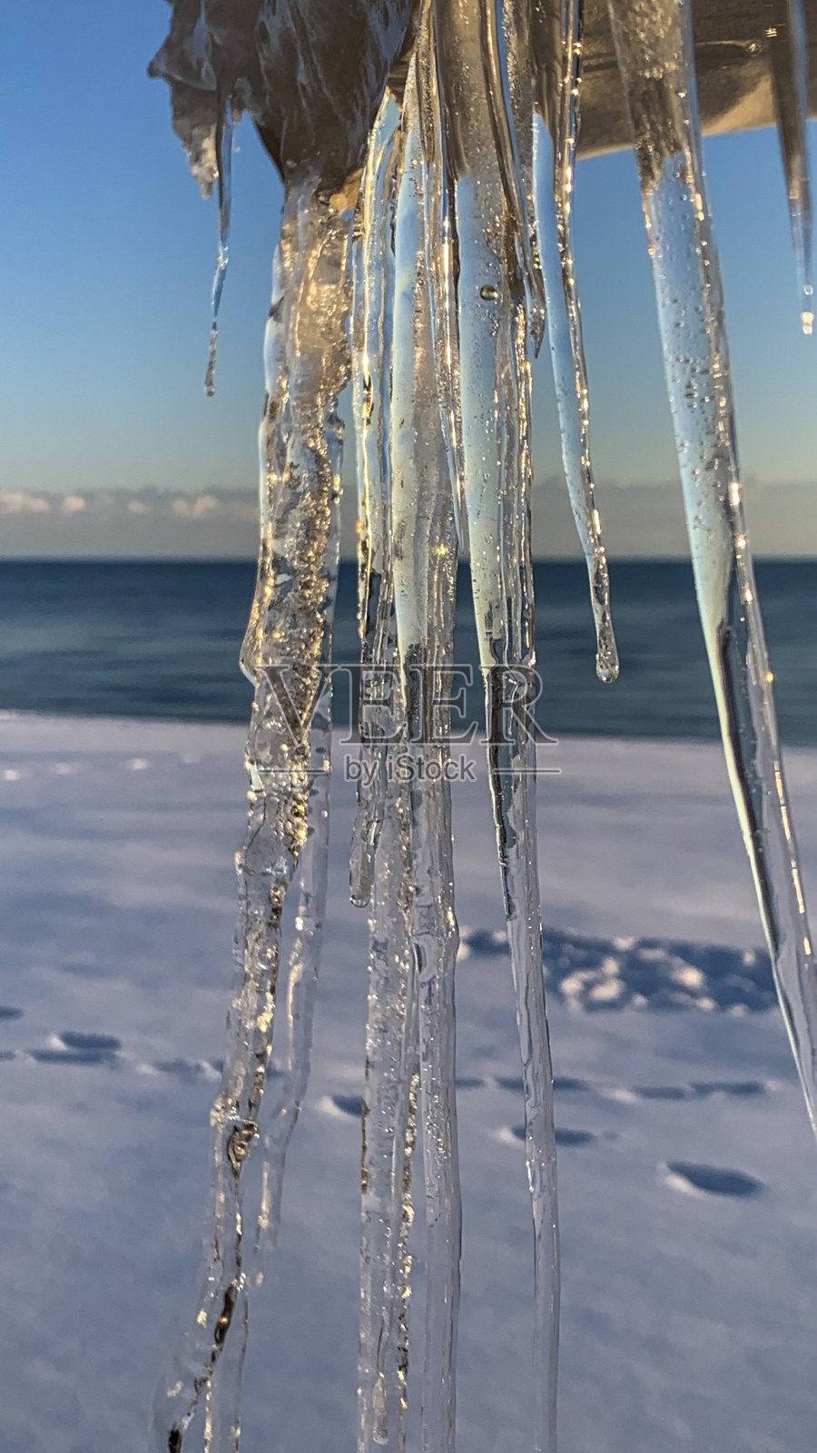 透明的冰柱在特写中悬挂下来，阳光反射在大海上，雪海滩上有脚步声照片摄影图片