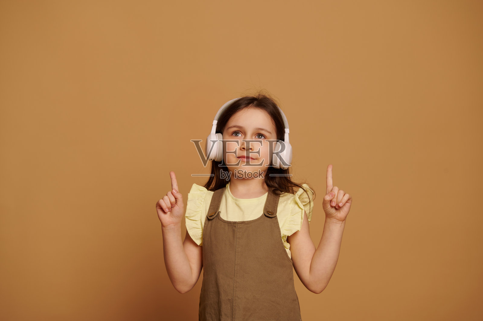 可爱的孩子女孩在无线耳机，指向她的食指在复制广告空间米色孤立的背景照片摄影图片
