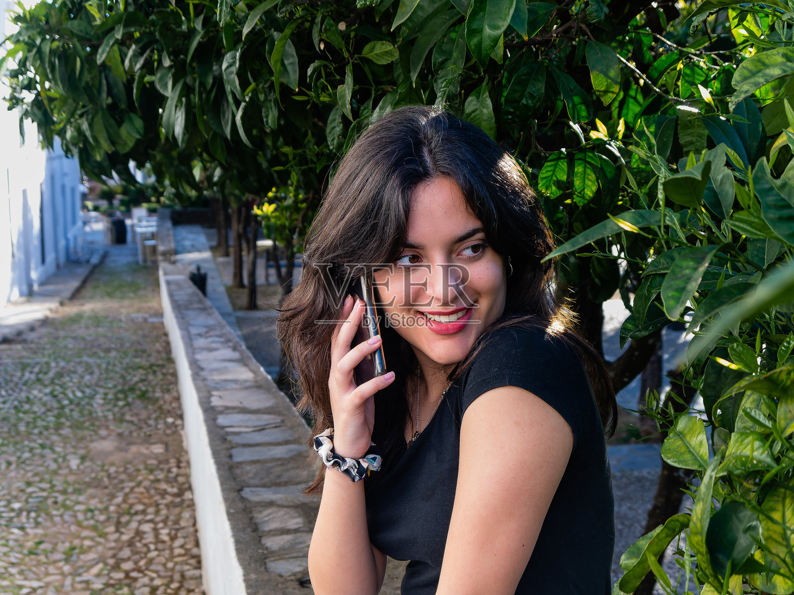 迷人的年轻女子，讲手机，坐在长凳与绿色树枝的背景，复制空间。照片摄影图片