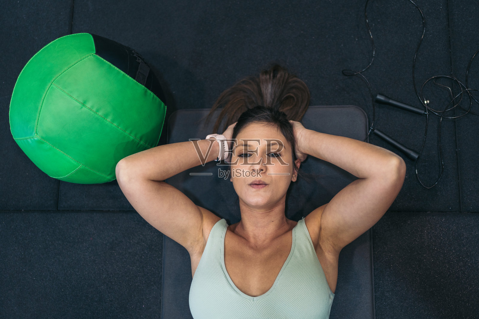 年轻的女运动员专注地躺在运动垫上，双手交叉放在脖子后面，旁边是一个软壁球和跳绳。照片摄影图片