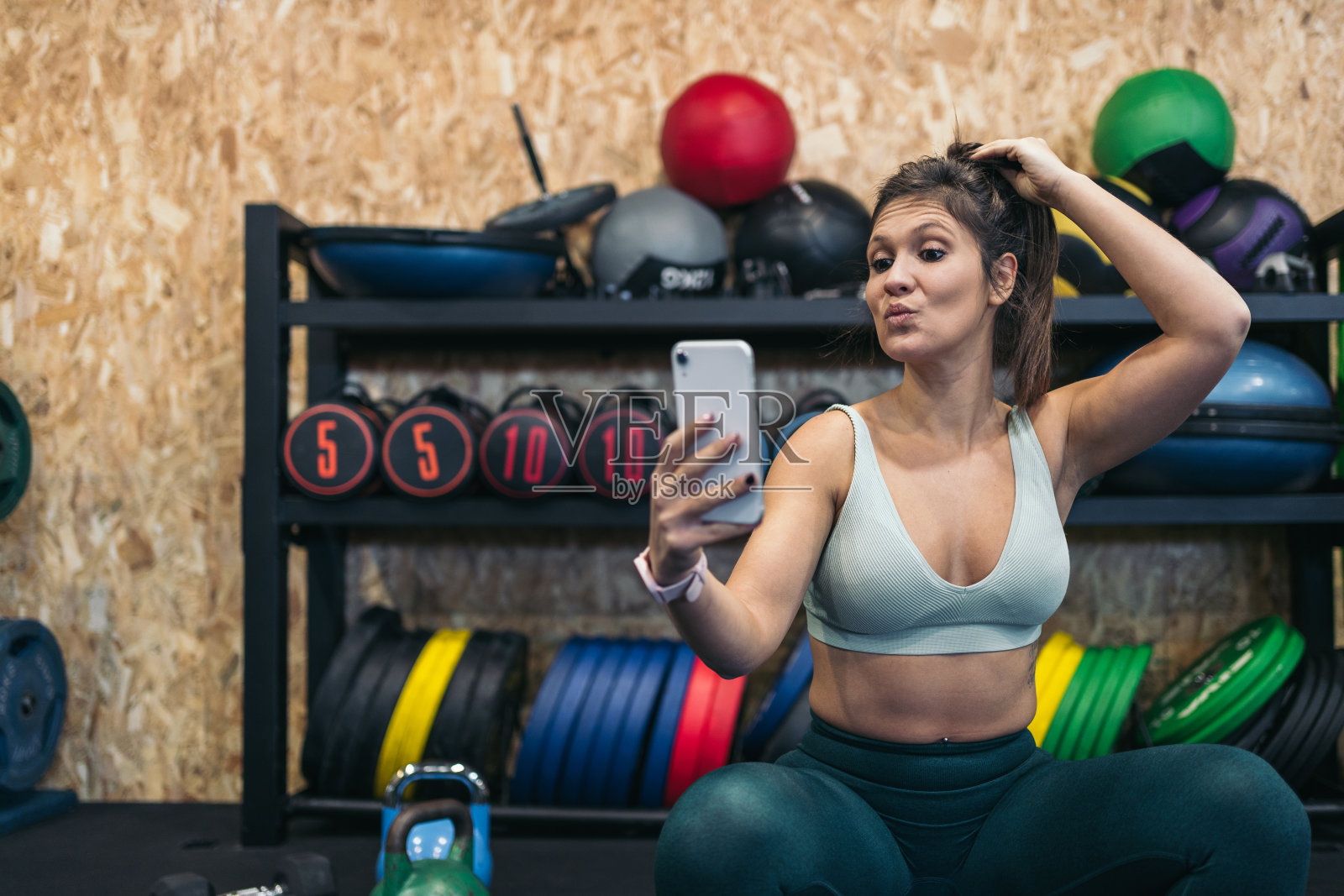 年轻的运动女性在健身房坐在一个柔软的壁球上抚摸她的头发，吹一个吻，在一次伟大而艰苦的锻炼后用手机自拍。照片摄影图片