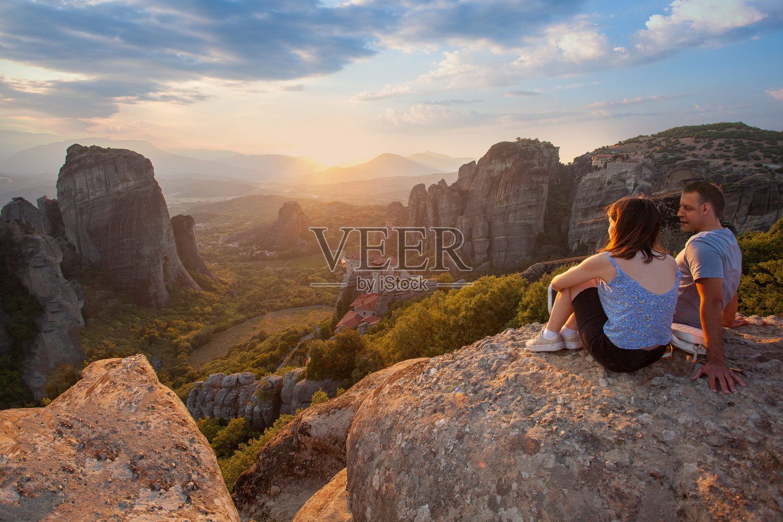 一对恋人坐在岩石上俯瞰夕阳下的迈泰奥拉。去希腊旅游。照片摄影图片
