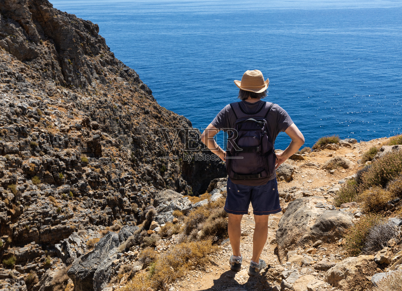 希腊克里特岛，一个戴着帽子的长发男子站在湛蓝大海的陡峭岸边照片摄影图片