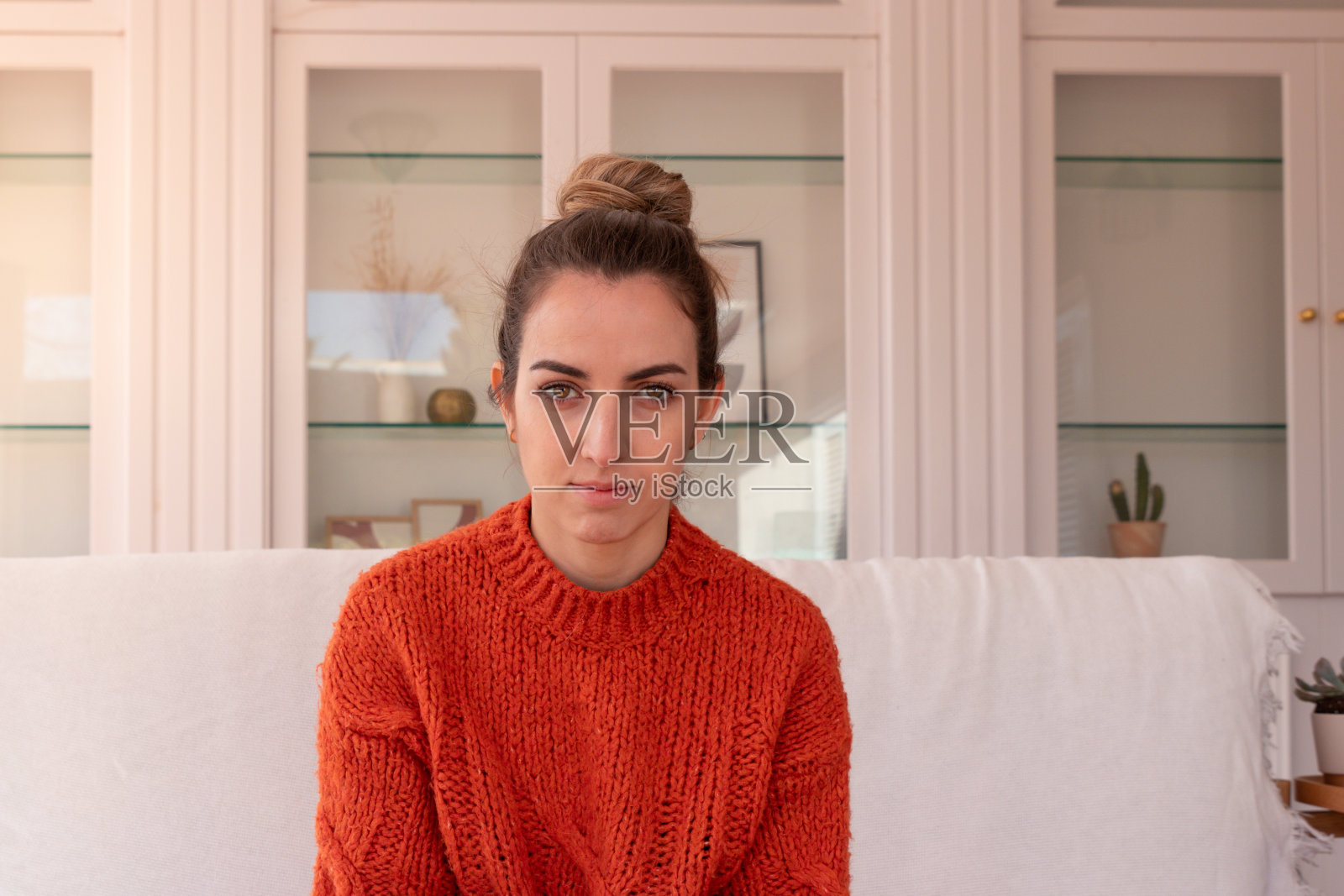 年轻女子与深色头发穿着温暖的橙色毛衣坐在白色沙发在家里看相机照片摄影图片