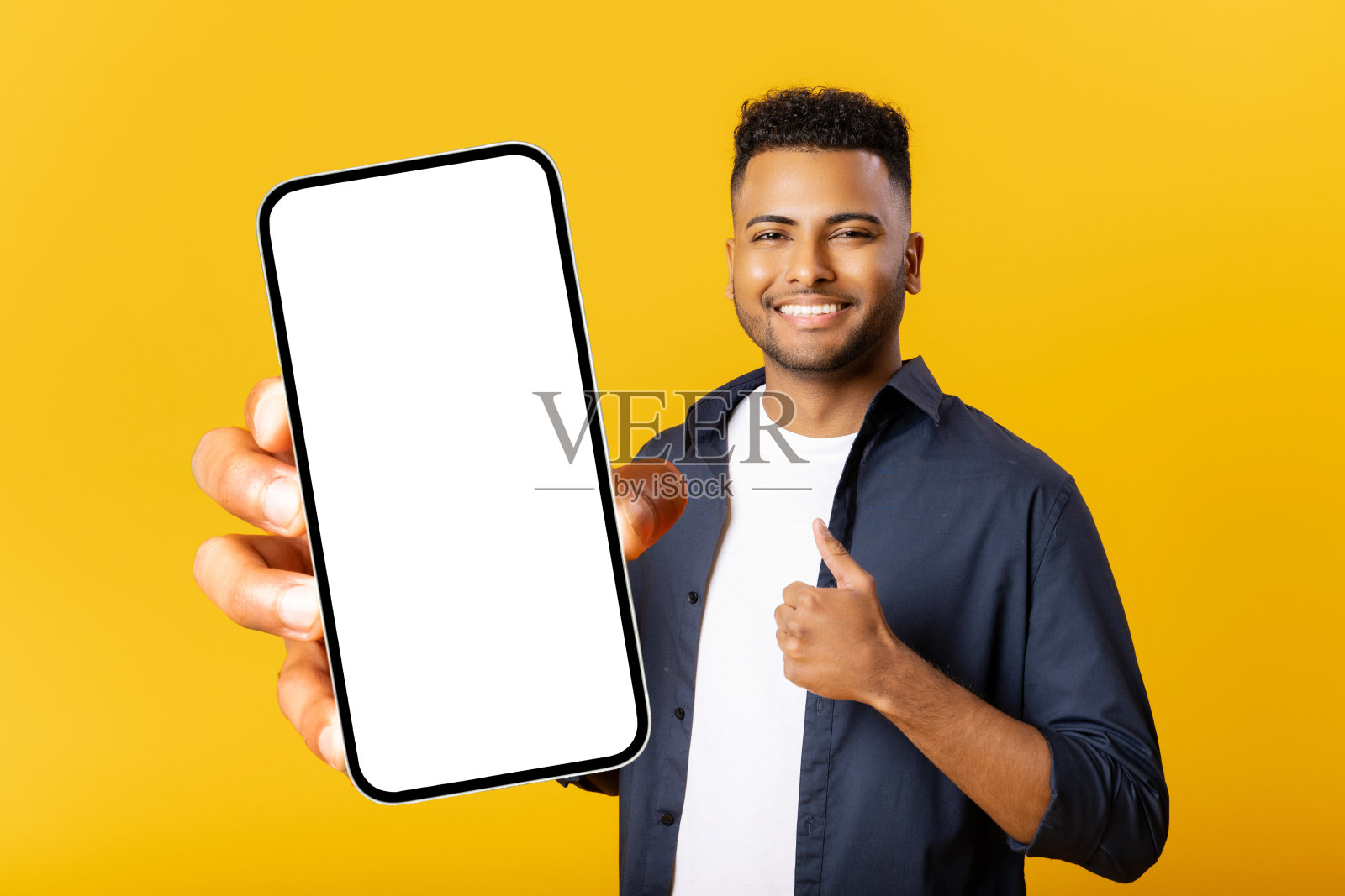 快乐的印度男子手里拿着智能手机，黄色背景，显示智能手机空屏，竖起大拇指，为手机应用做广告照片摄影图片