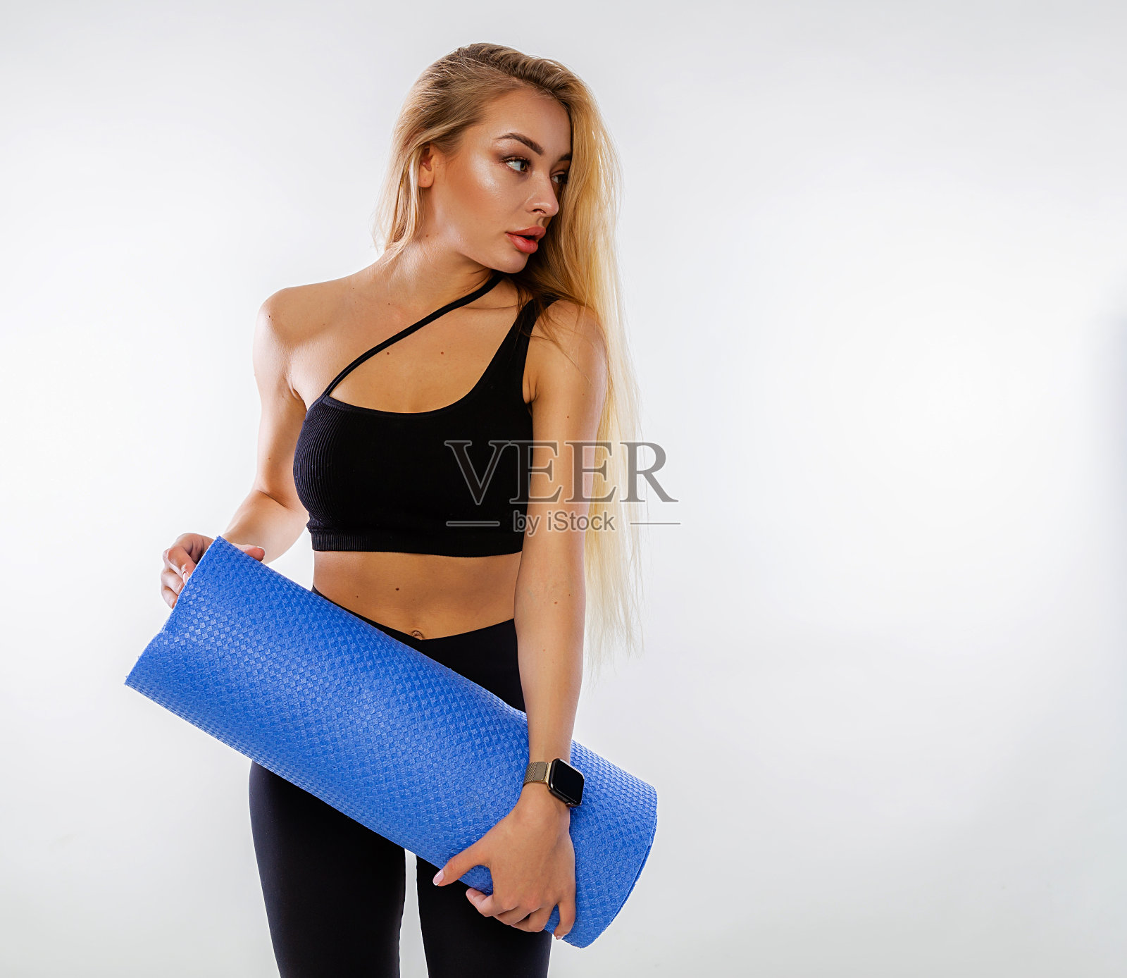 身穿黑色运动服、金黄色长发的白人女性在白色背景上举着瑜伽垫或健身垫摆姿势，展示着完美的身材。个人护理和在家运动。照片摄影图片