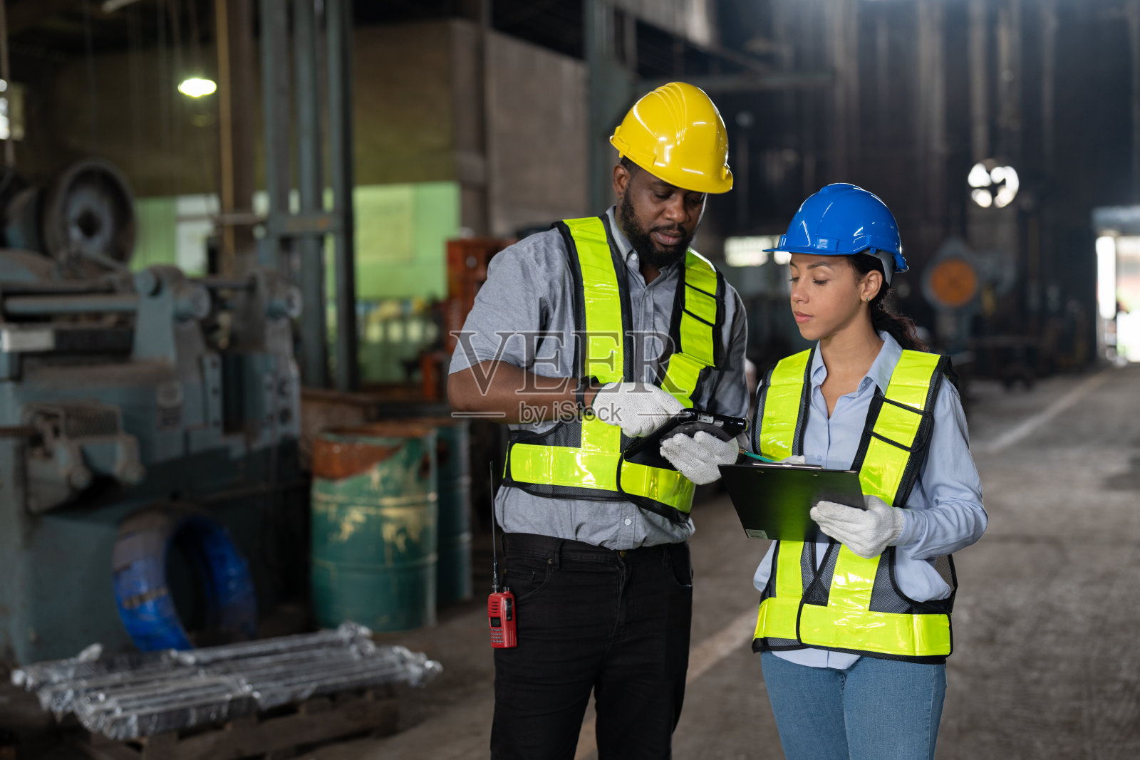 身穿安全背心和头盔的非裔美国男女工程师在重金属工业工厂工作。两名技术领班在工厂讨论检修机器的问题。照片摄影图片