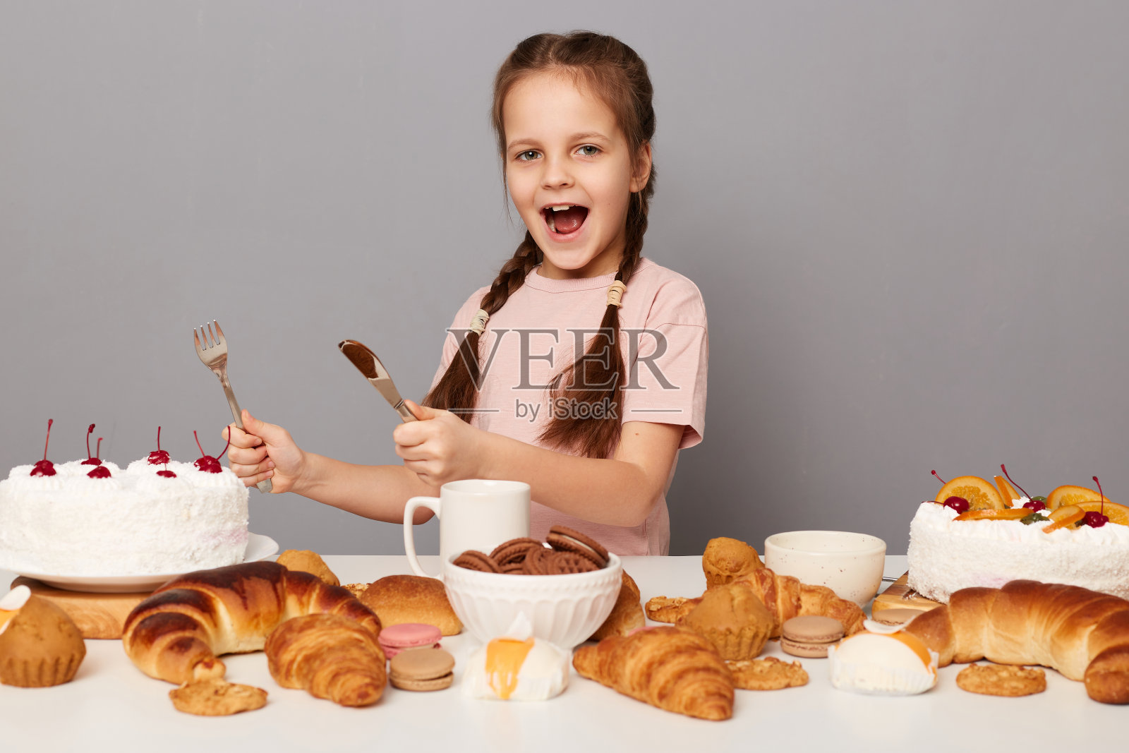 兴奋饥饿的疯狂的小女孩辫子摆姿势靠近桌子与美味的糖果，拿着刀叉准备吃生日蛋糕孤立在灰色背景的肖像照片摄影图片