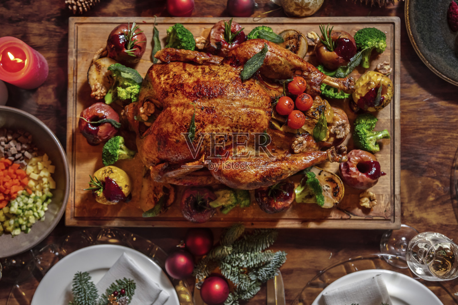 塞满西兰花、苹果、迷迭香、核桃、西红柿的炸鸡放在木板上，周围是喜庆的桌子、盘子、杯子和圣诞树装饰品照片摄影图片
