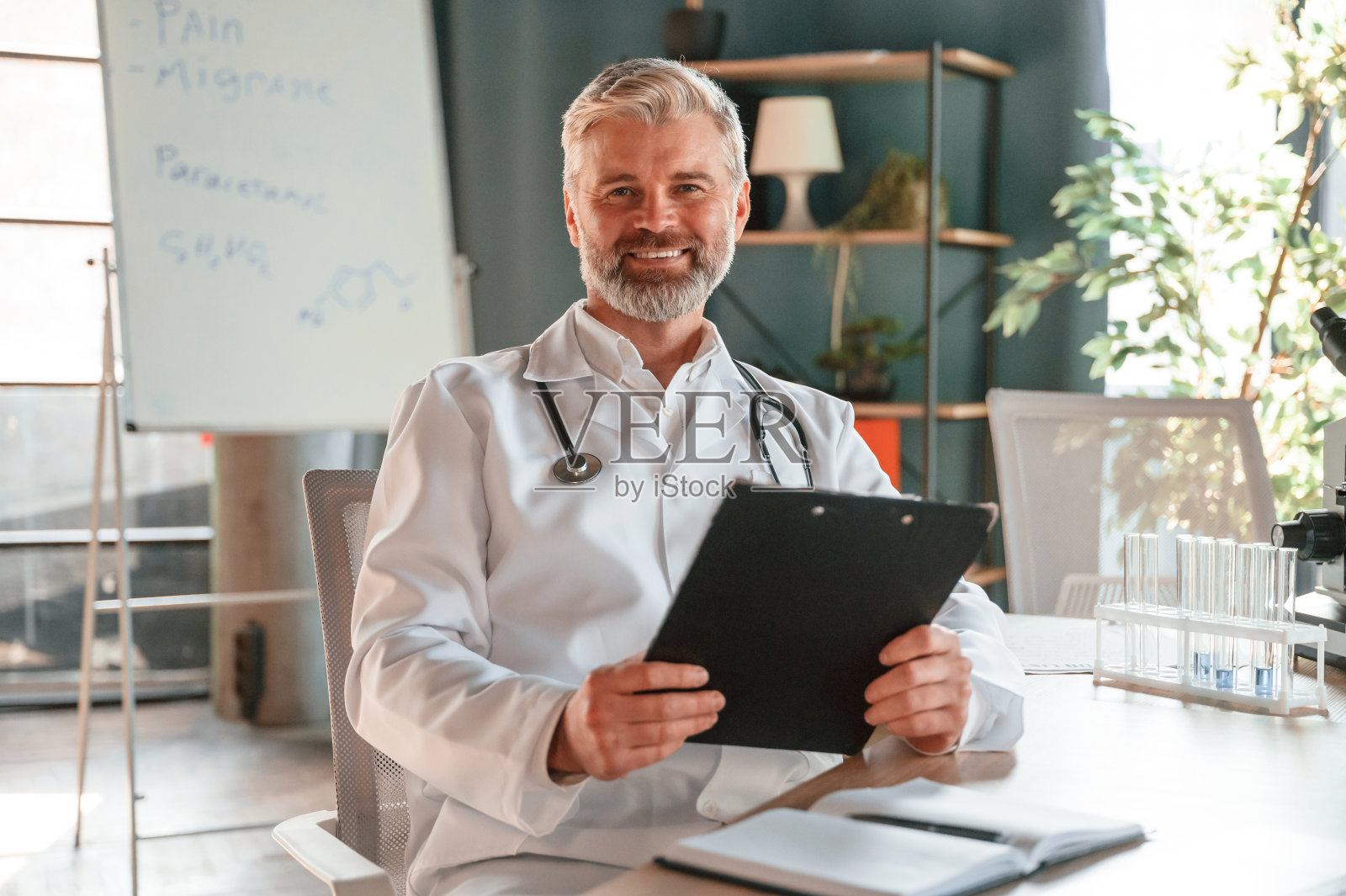 坐着微笑。穿白大褂的资深医生正在室内工作照片摄影图片