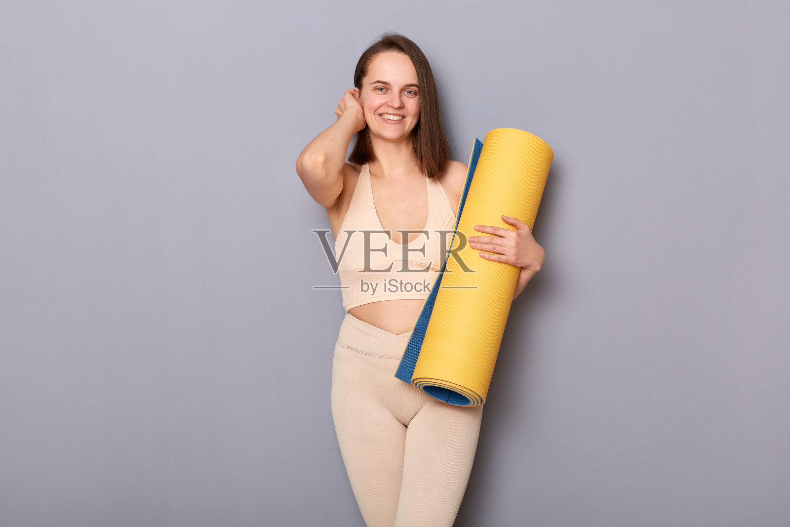 美丽迷人的运动女性抱着卷起的健身垫，看着相机，很高兴在健身房做运动，站在灰色背景孤立的肖像。照片摄影图片