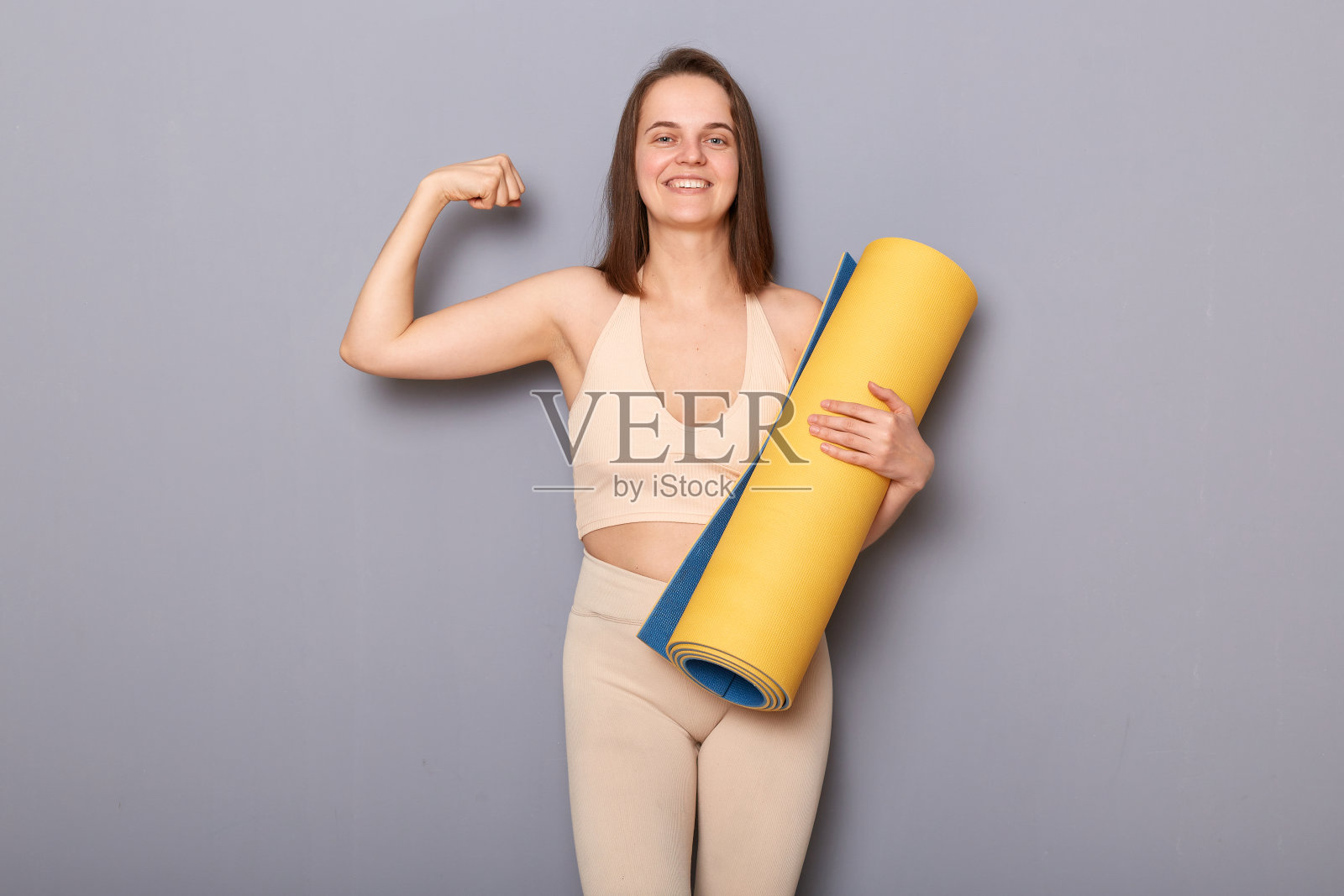 肖像满意自豪的运动员在米色运动服抱着健身垫，展示她的肱二头肌，在运动训练后展示她的力量。照片摄影图片