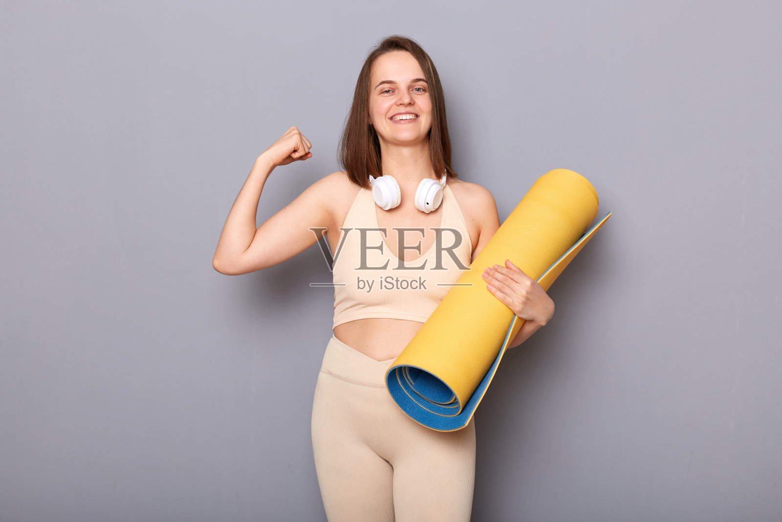 满意的高兴的女人的肖像棕色头发站在瑜伽垫和耳机在脖子上，显示她的肱二头肌，微笑，享受锻炼和减肥的结果。照片摄影图片