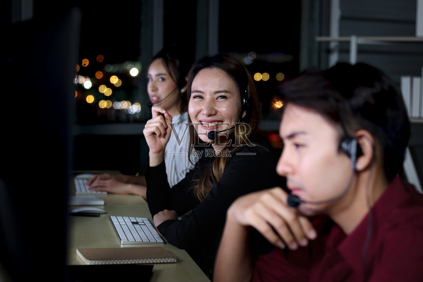 快乐微笑的亚洲女性在呼叫中心工作，与队友同事一起通宵工作，夜班客服人员戴着麦克风耳机使用电脑为客户提供支持照片摄影图片