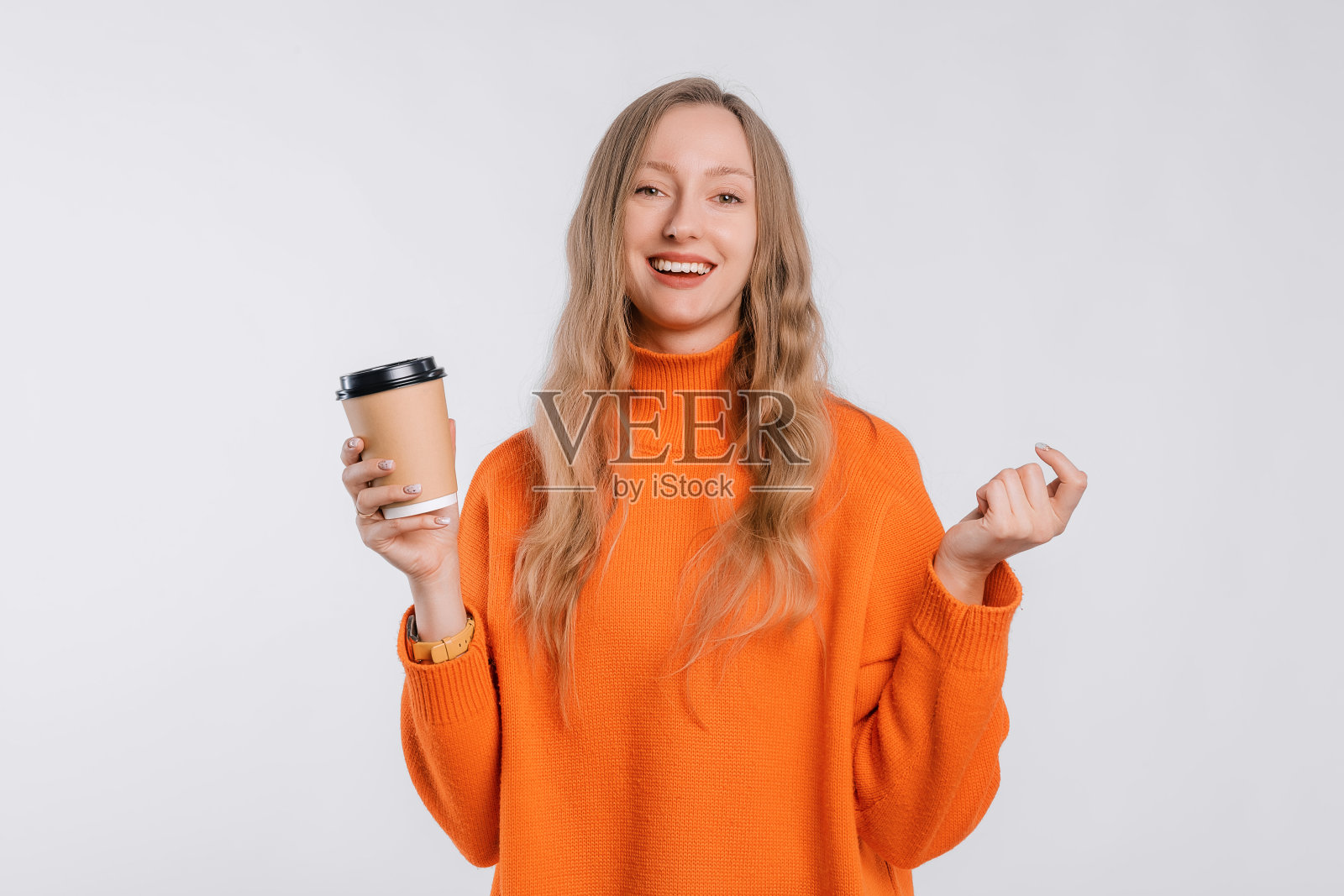 肖像一个快乐的年轻女子在舒适的橙色毛衣拿着纸杯咖啡，站在中性的背景照片摄影图片