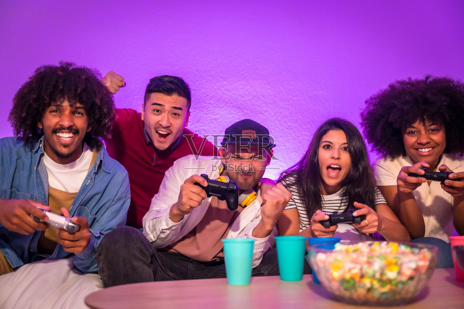 成人派对。有魅力的年轻人坐在沙发上，拿着爆米花玩电子游戏。用操纵杆或控制器在手，看着显示器照片摄影图片