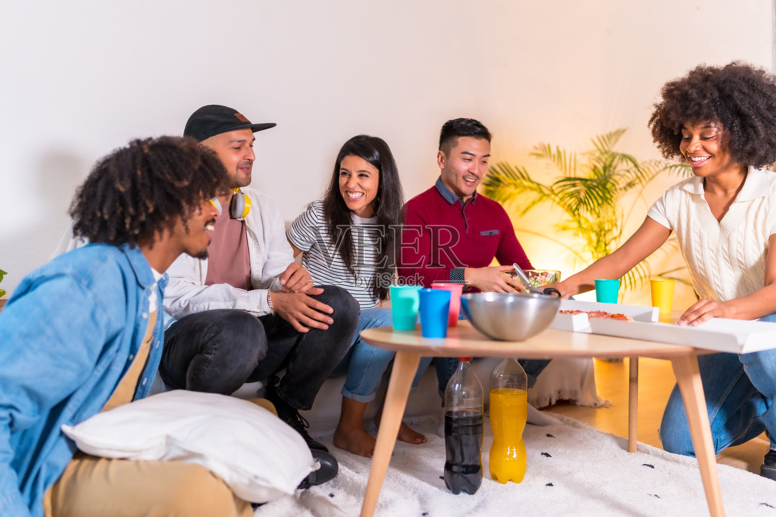 在一个家庭聚会上，一群多民族的朋友坐在沙发上吃着披萨，喝着软饮料，一个年轻人从街上带着披萨盒来了照片摄影图片