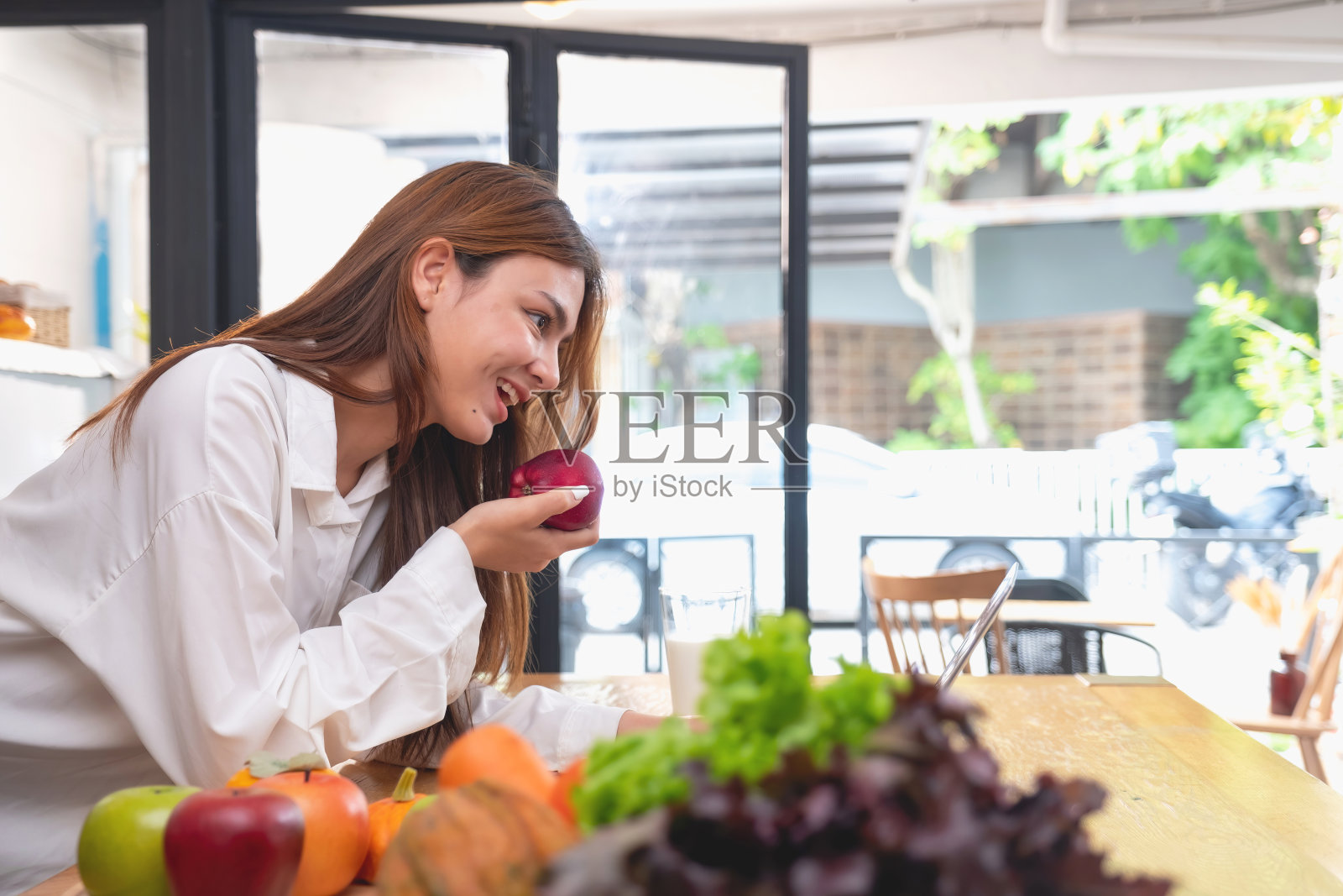 一位穿着白衬衫的美女正在厨房里做健康的早餐，有面包、蔬菜、水果和牛奶，并打开笔记本电脑上烹饪课。健康的烹饪理念。照片摄影图片