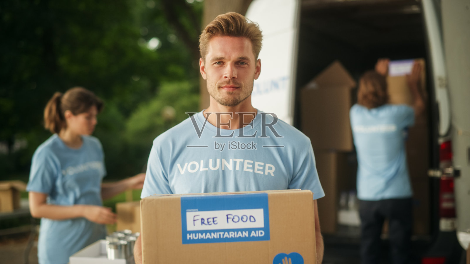 一个快乐的年轻男性志愿者的肖像。穿着蓝色t恤的英俊白人男子微笑着向观众伸出人道主义援助箱。捐赠中心和志愿服务概念。照片摄影图片