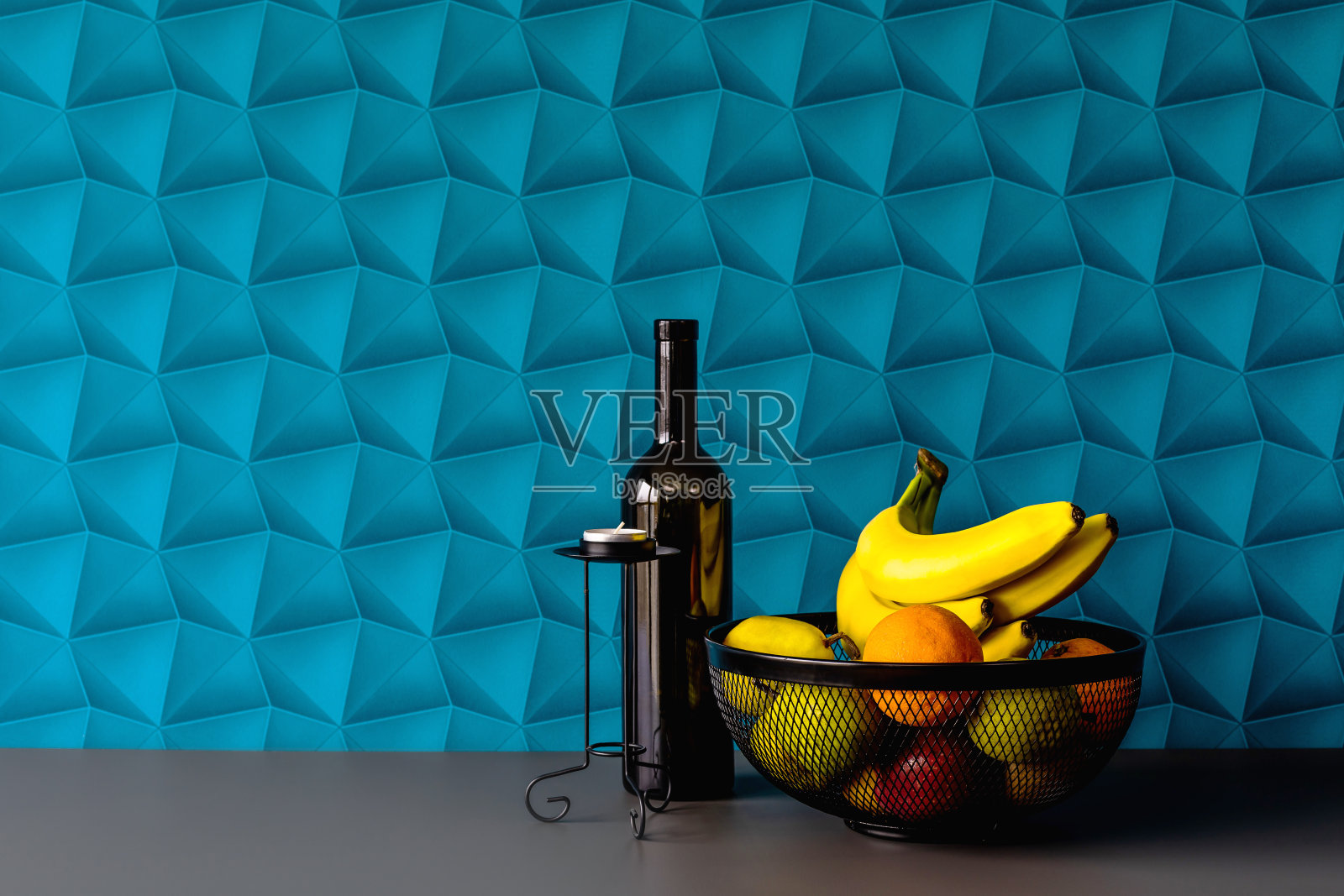 背景是蓝色的壁纸——色彩丰富的空间。黑色水果篮的优雅排列。黑色烛台与蜡烛暖器，黑色酒瓶作为装饰。新鲜水果，如香蕉、橙子、梨和苹果。照片摄影图片