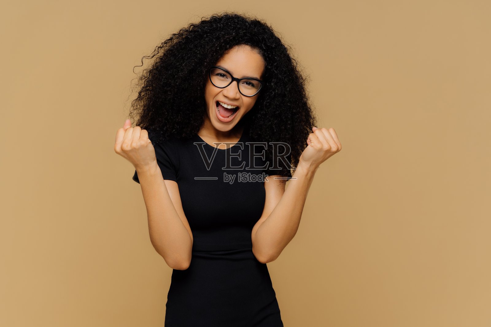 胜利可爱的黑皮肤女人举起紧握的拳头，欢呼雀跃，庆祝胜利，穿着黑色的衣服，站在棕色的背景，感觉像赢家。肢体语言，成功理念照片摄影图片