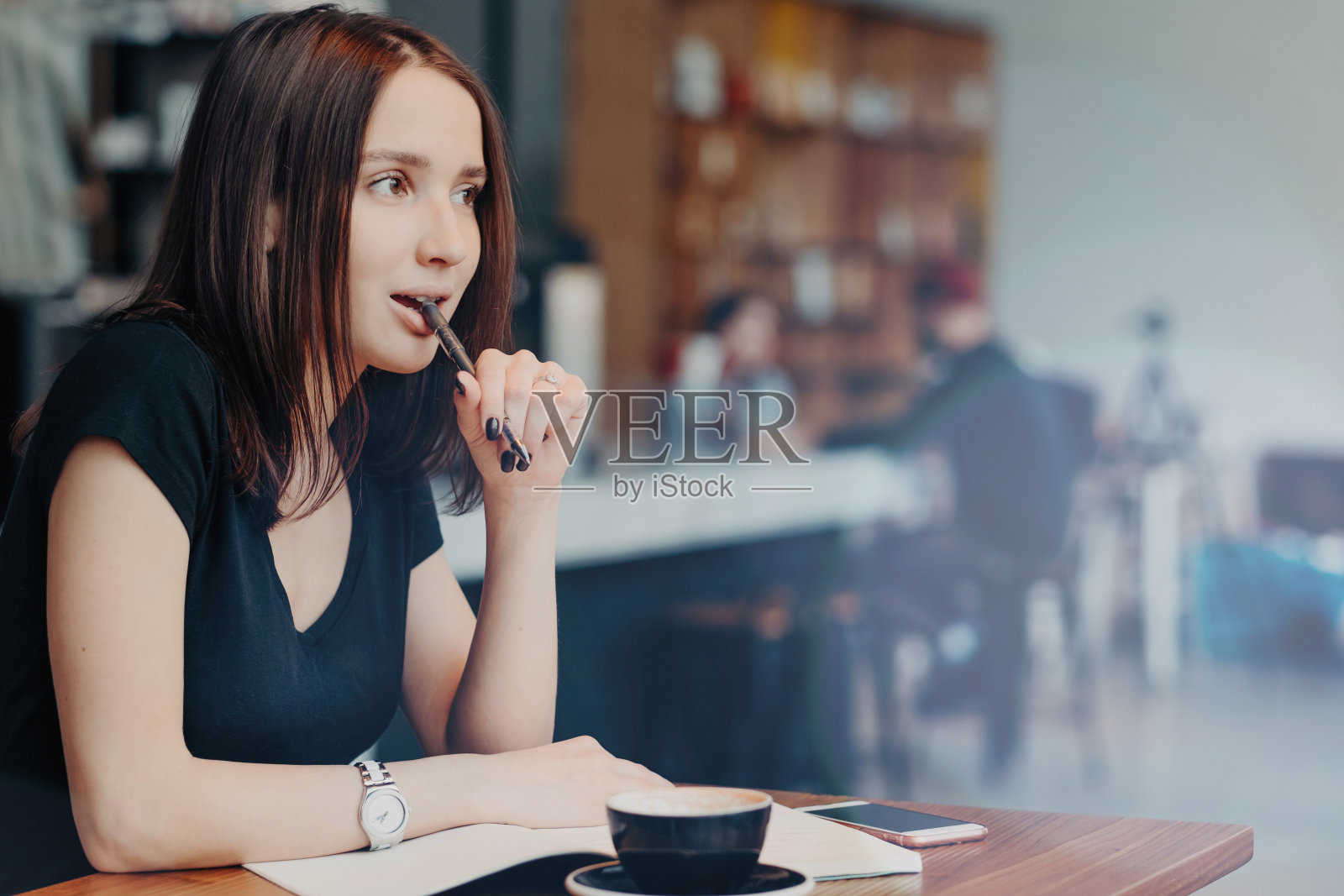 沉思的女博主在记事本上写下发表在博客上的想法，坐在咖啡馆里，把笔放在嘴里，专注地喝着咖啡，穿着黑色t恤，思考着计划照片摄影图片