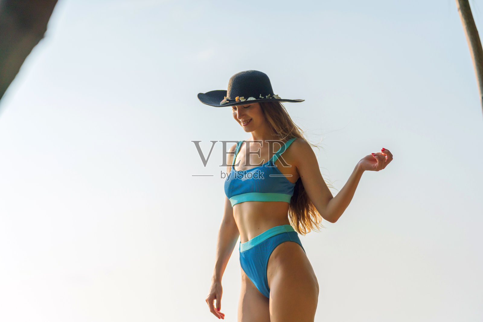 生活是一片海滩。美丽的白人女子在蓝色比基尼黑色沙滩帽和太阳镜漫步在海滩上享受海风和太阳晒黑照片摄影图片