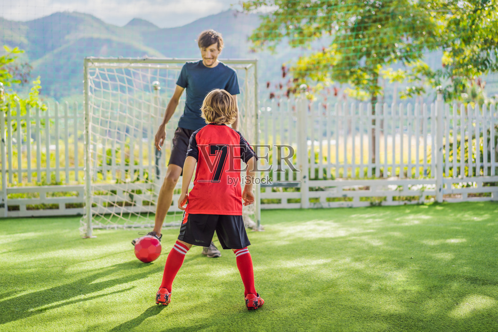 穿着红色足球服的小男孩和他的教练或父亲在户外踢足球。活泼的孩子与孩子或父亲一起运动，微笑的快乐男孩在夏天玩得开心照片摄影图片