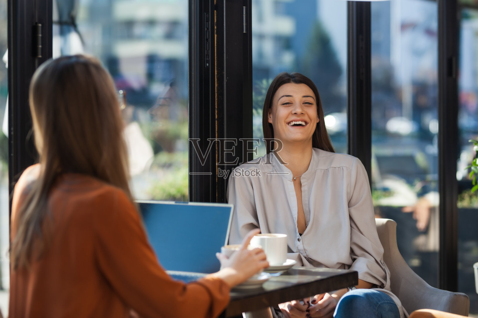 两个年轻的女商人在咖啡馆里一对一地开会。下班后的朋友们在阳光明媚的日子里闲聊，坐在靠窗的桌子上喝咖啡。照片摄影图片