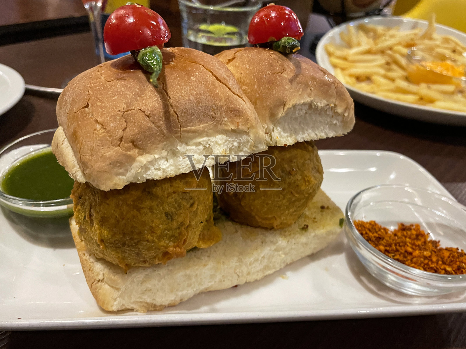 餐厅场景的特写图像，盘子里有孟买vada pav，油炸土豆饺子在面包包上，上面有青椒和番茄，重点在前景照片摄影图片