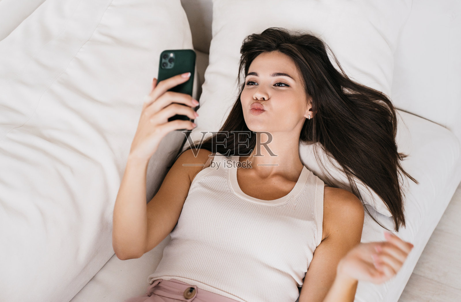 俏皮的亚洲年轻女子在休闲装躺在沙发上视频通话使用电话发送飞吻看着屏幕。年轻的韩国女子周末在家放松与男友谈话。照片摄影图片