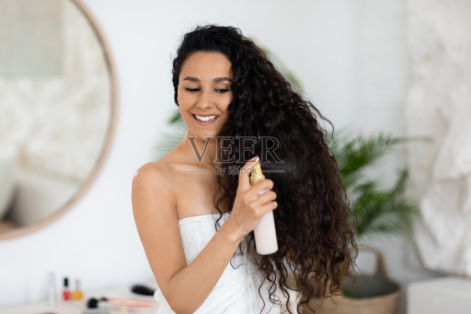 快乐的千禧一代白人女士用毛巾裹着，在长长的卷发上喷洒护理液，享受护理照片摄影图片