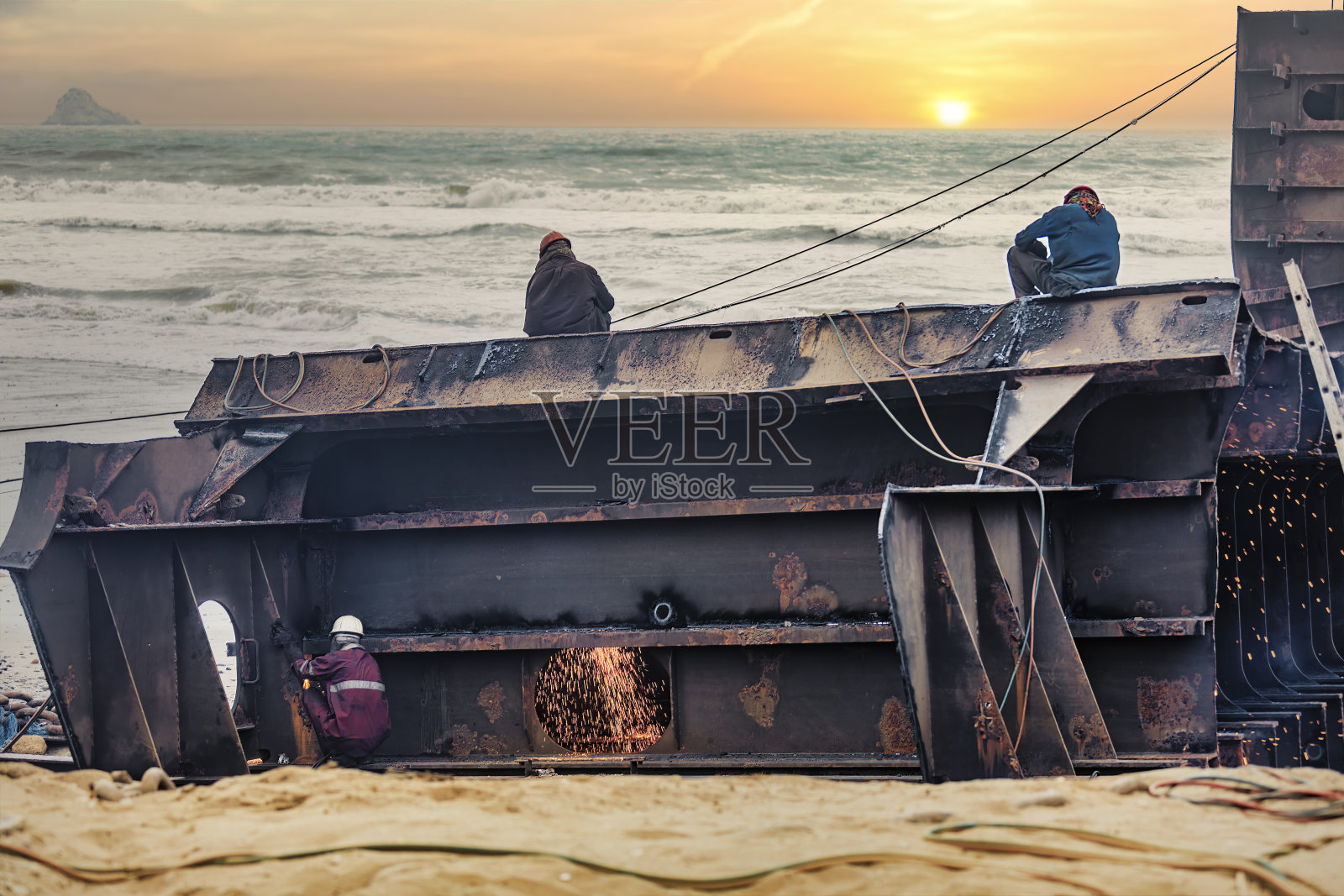 一艘大型油轮在港口拆船，工人通过焊接切割零件，背景是巴基斯坦加达尼拆船场海滩上的日落照片摄影图片