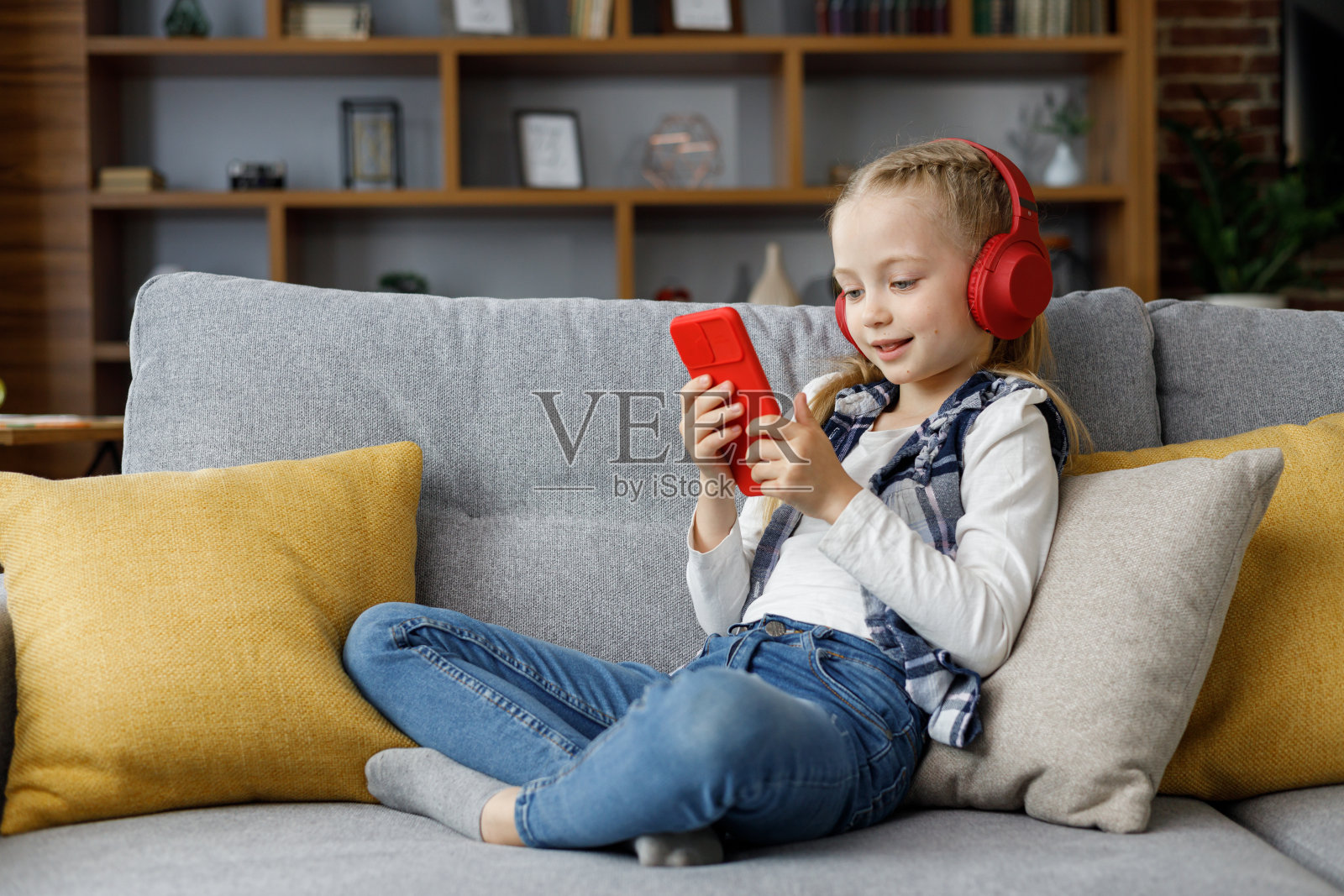 可爱的小女孩戴着耳机躺在家里的沙发上。选择最喜欢的音乐或动画片，发短信，上网，看视频，玩智能手机游戏。照片摄影图片