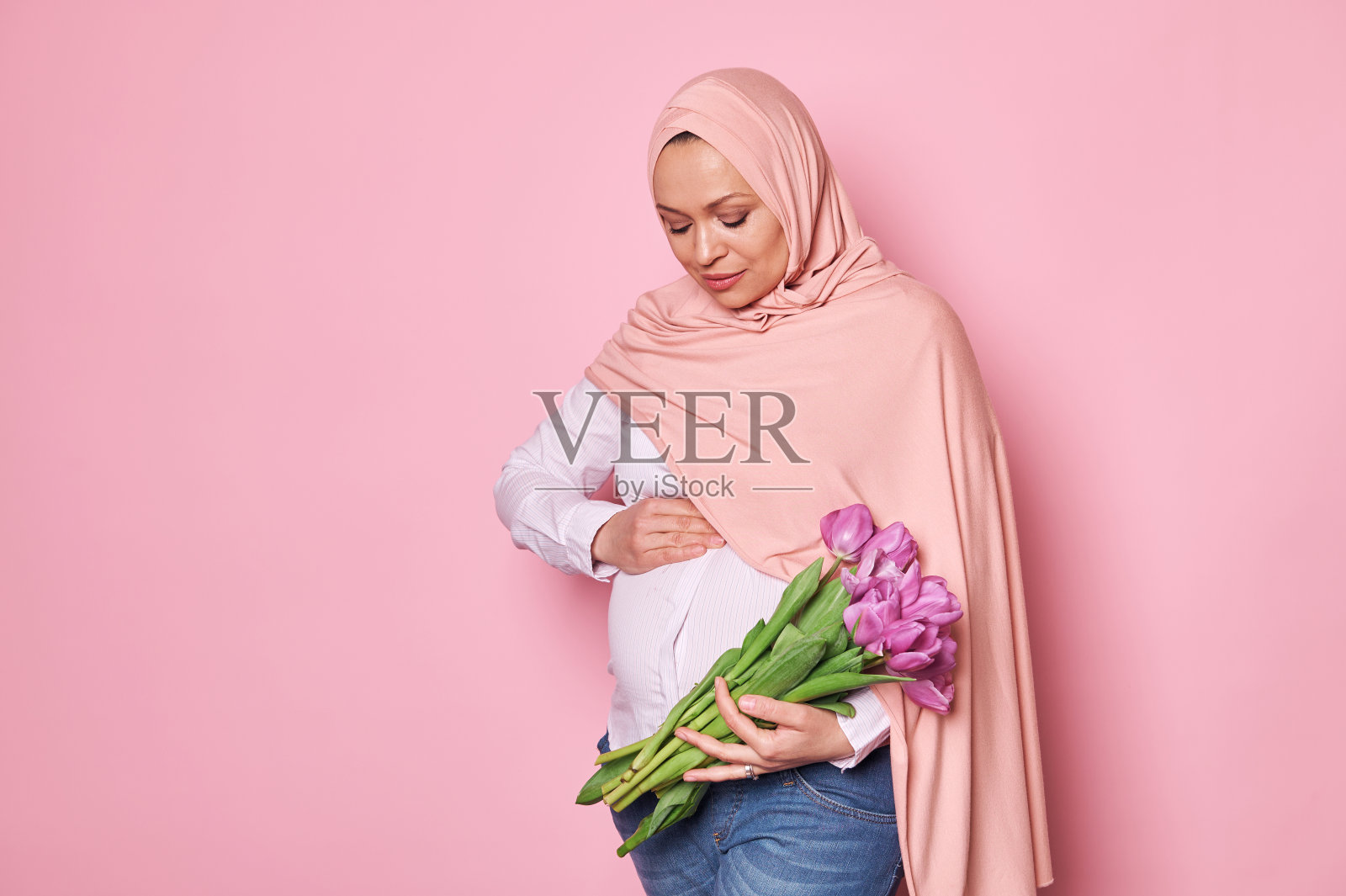 戴着粉色头巾的穆斯林孕妇，摸着她的肚子，感受着婴儿的第一次踢腿，拿着美丽的郁金香，孤立在粉色上照片摄影图片