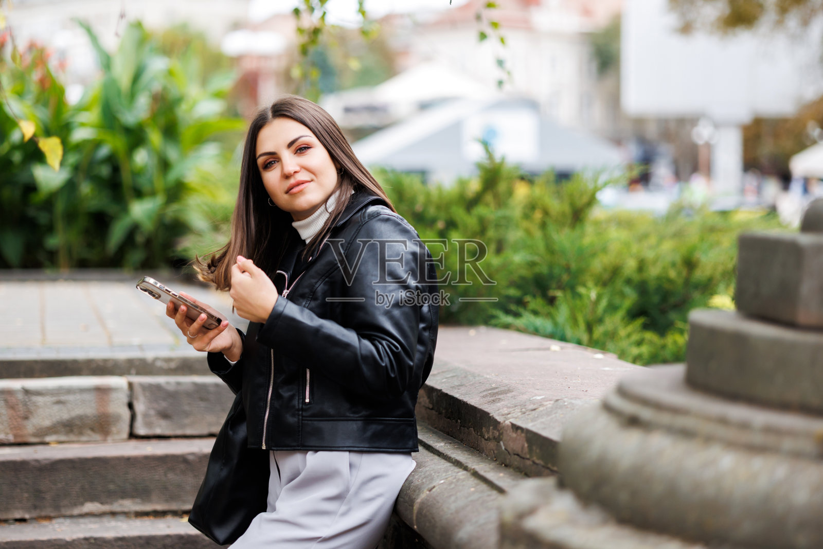 一个年轻漂亮的女孩穿着黑色皮夹克走在公园的街道上，用她的智能手机工作。自由职业者兼博客作者的作品。照片摄影图片