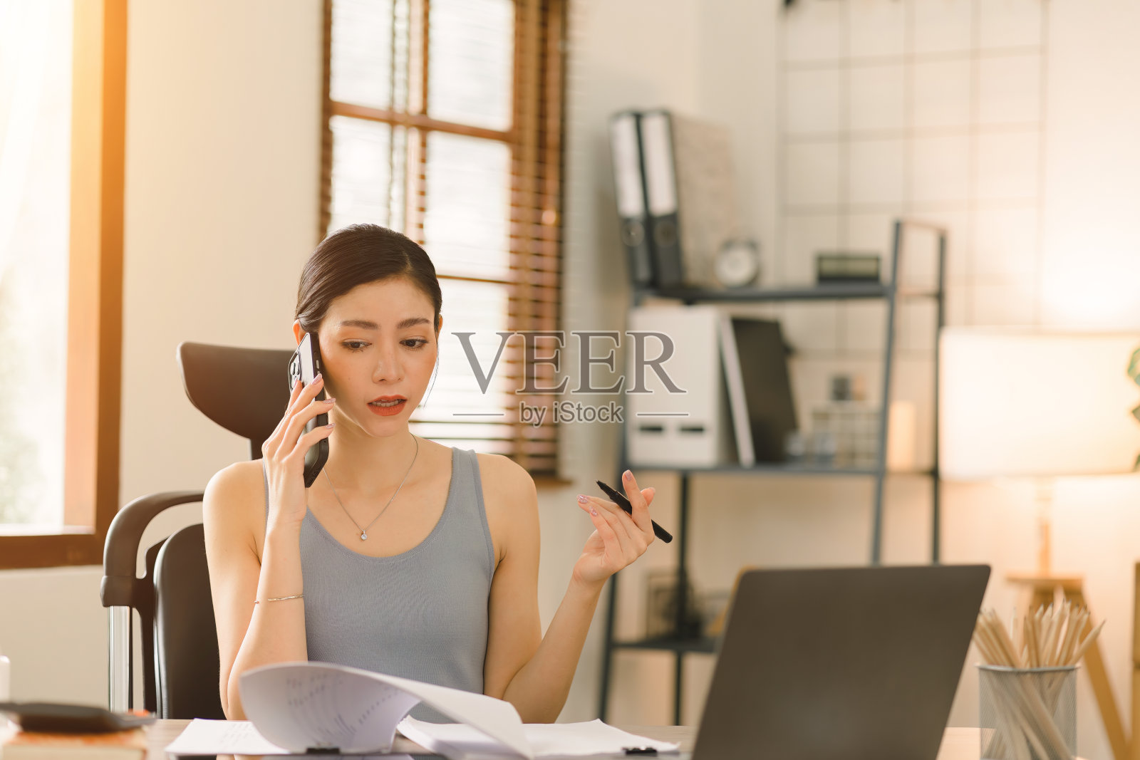 一位千禧一代的创业女商人坐在家里，一边用智能手机打电话，一边用笔记本电脑工作，一边通过家庭办公室管理自己的业务。温暖的色调照片摄影图片