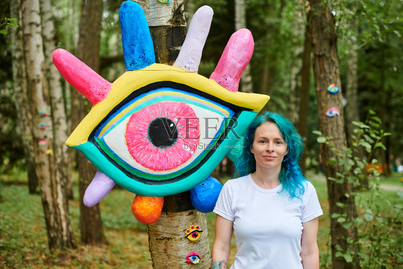 在大眼艺术装置旁，穿着白色T恤，染成蓝绿色头发的微笑年轻成年女子照片摄影图片